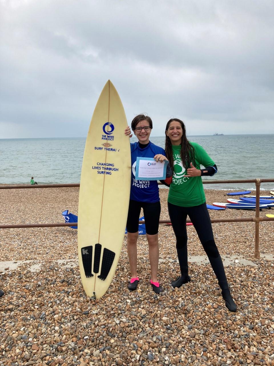 2 women on a pebble beach with sea and sky in the background. One is holding a surfboard and a certificate.