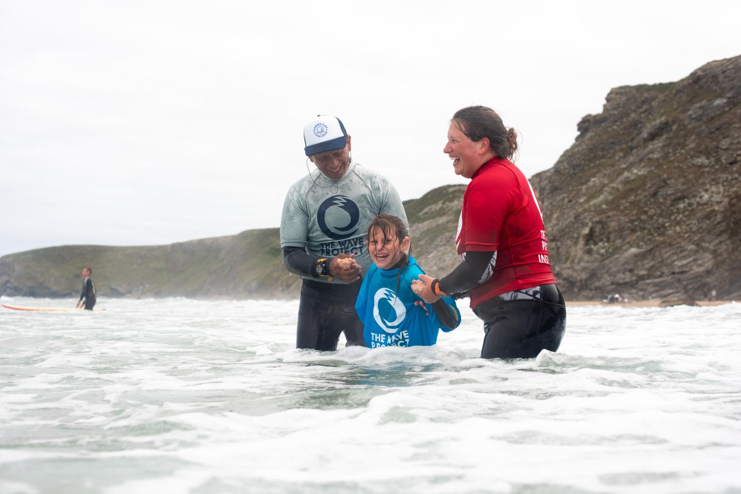 2 adults on each side of a child, holding her hands in the sea with cliff behind them