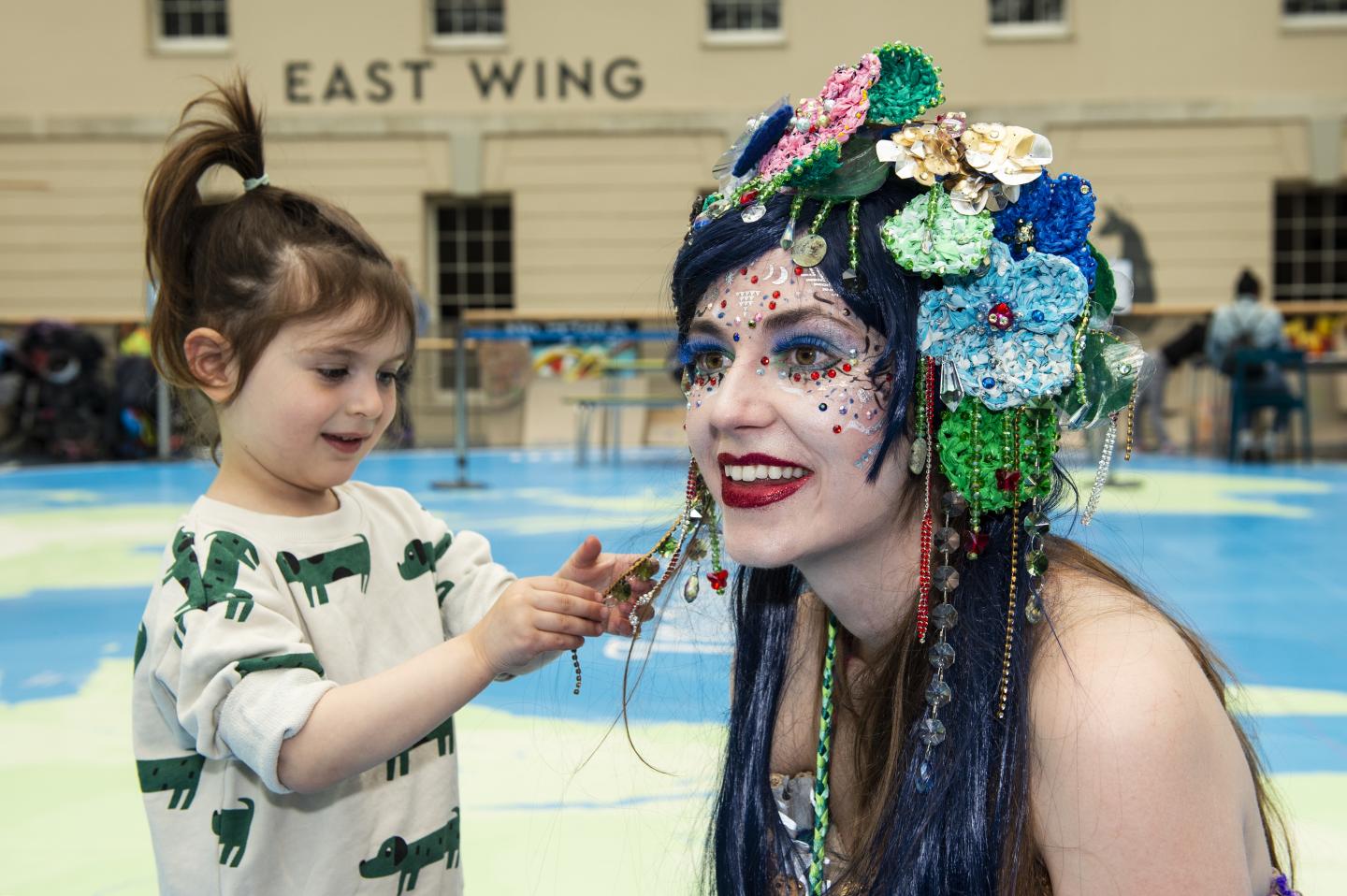 The image shows a child playing with the hair of a woman dressed up as a mermaid.