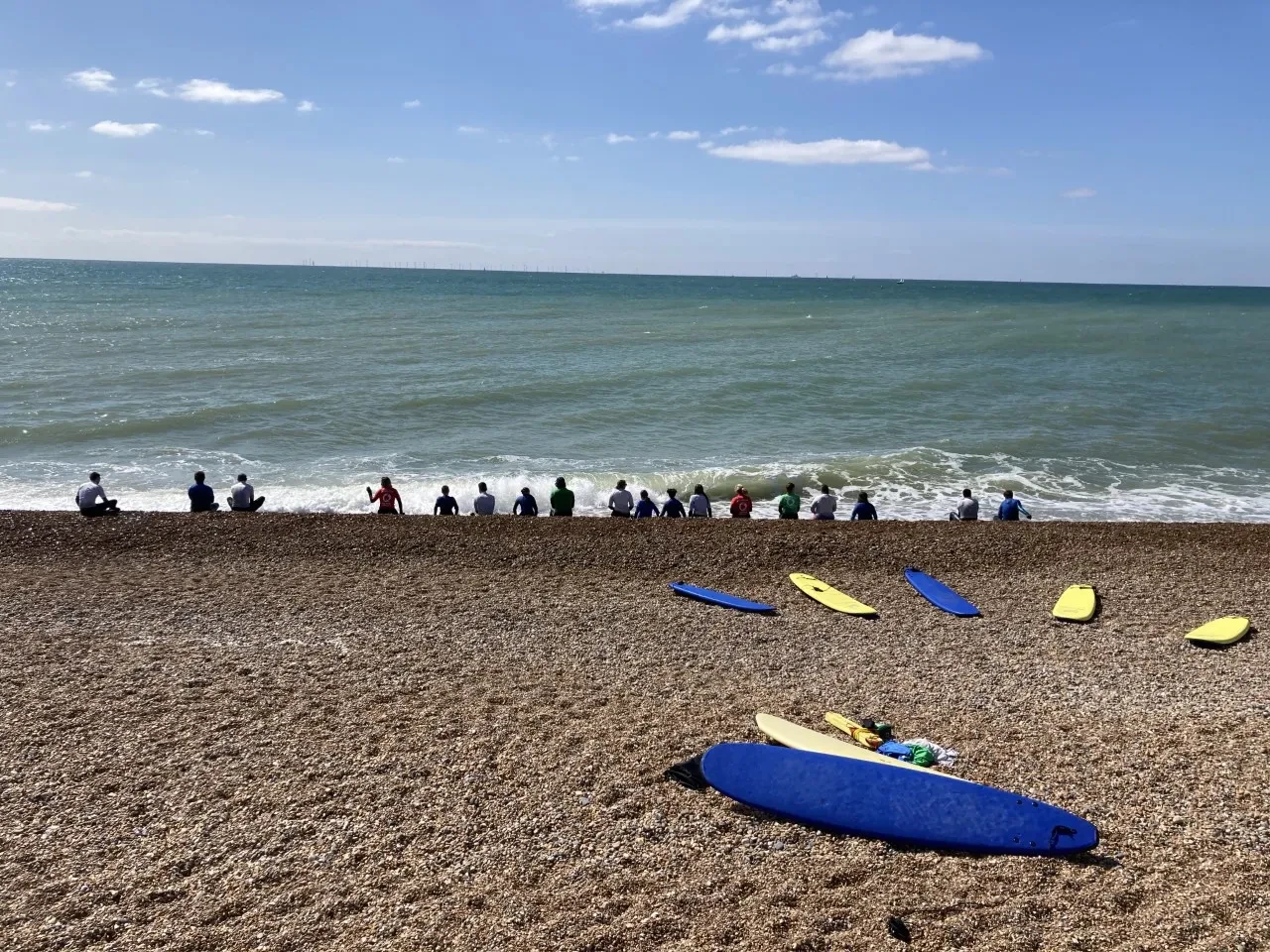 A beach with some surfboards in the foreground, a row of people with their backs to the camera, sitting on the beach looking out to the sea and horizon.