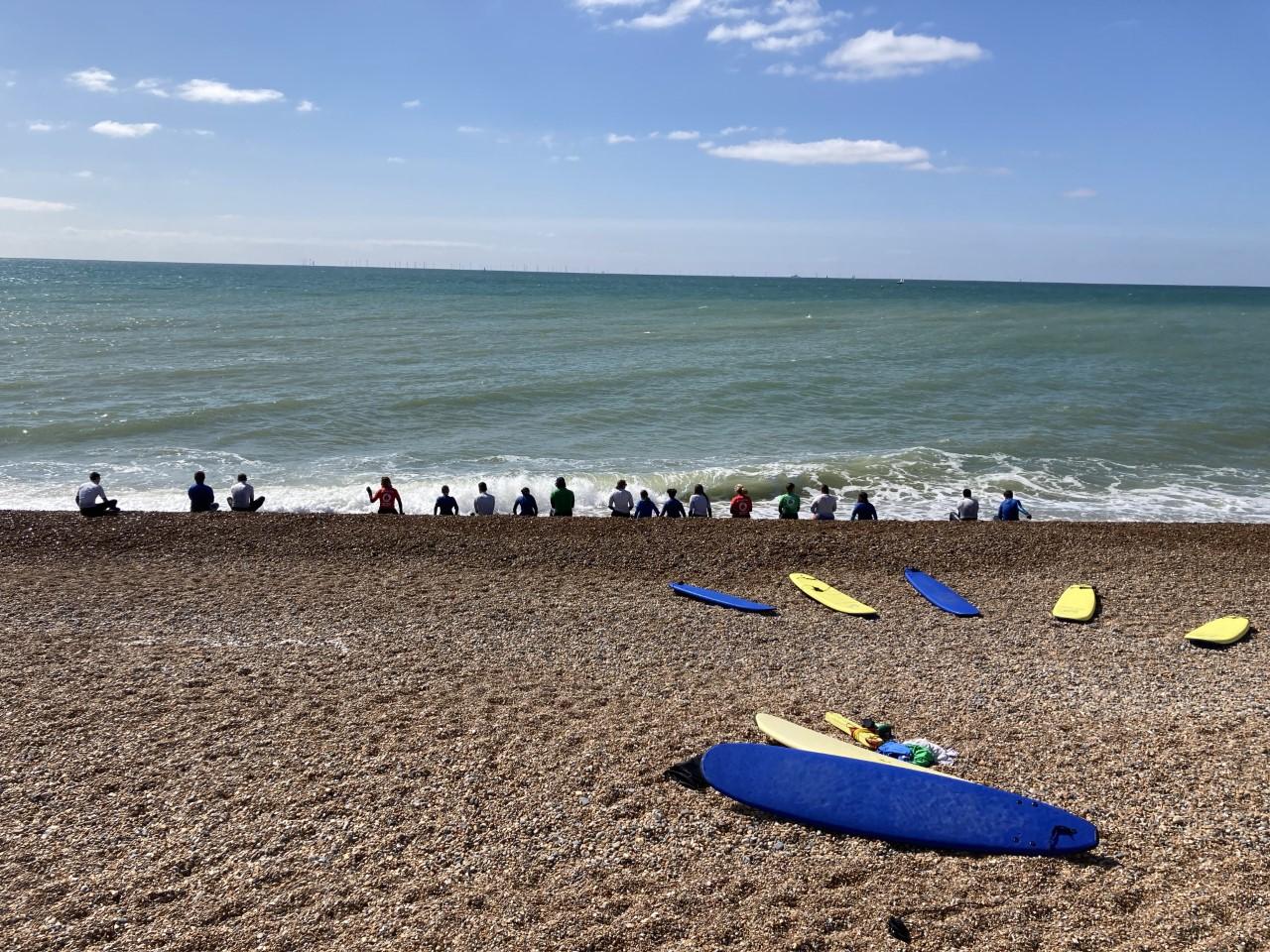 A beach with some surfboards in the foreground, a row of people with their backs to the camera, sitting on the beach looking out to the sea and horizon.