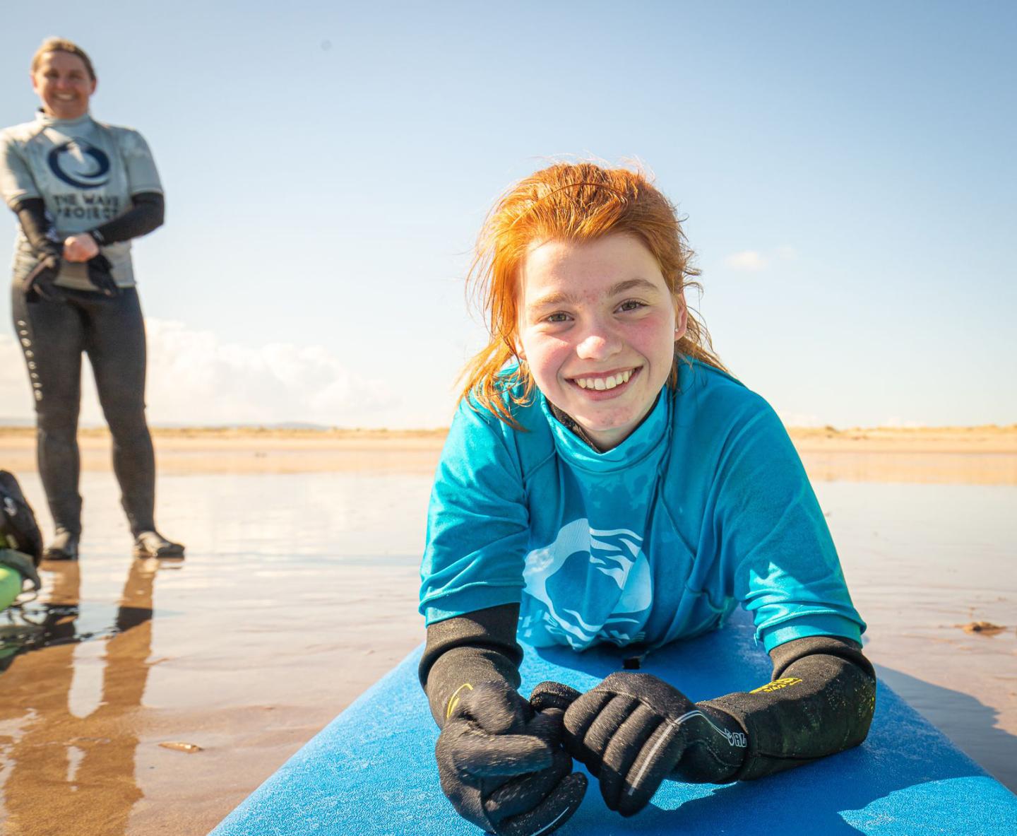 A girl lying on a surfboard on the beach with an adult watching on.