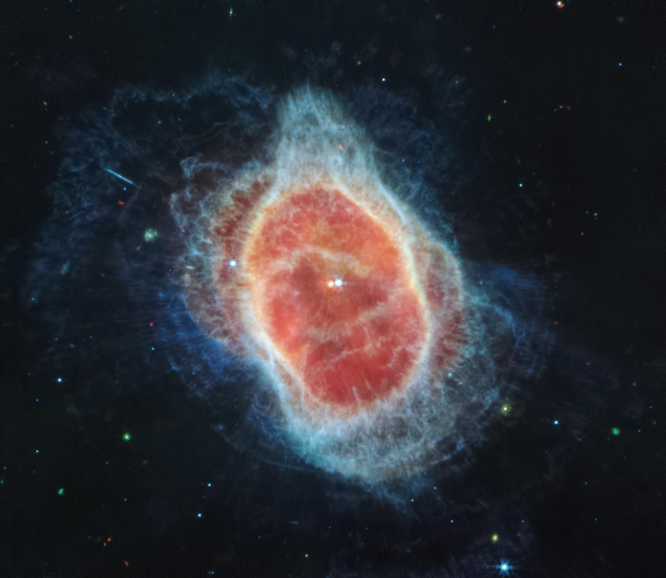 The Southern Ring Nebula is a large, semi-transparent oval that is slightly angled from top left to bottom right. Two stars appear at the center very close to one another. The one at left is red, the one at right is light blue. The blue star has tiny diffraction spikes around it. A large translucent red oval surrounds the central stars