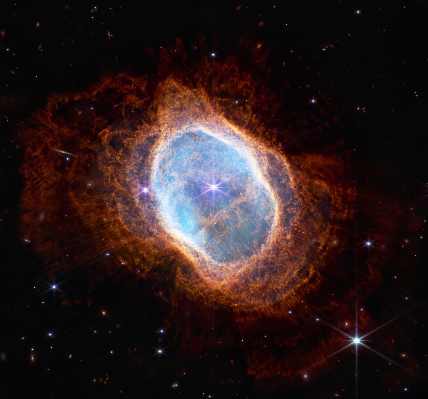 Colorful image of near-infrared light from a glowing cloud with a distorted ring-like shape, illuminated from within by a bright central star. The Southern Ring Nebula is a large, semi-transparent oval that is slightly angled from top left to bottom right. A bright white star appears at the center of this image.