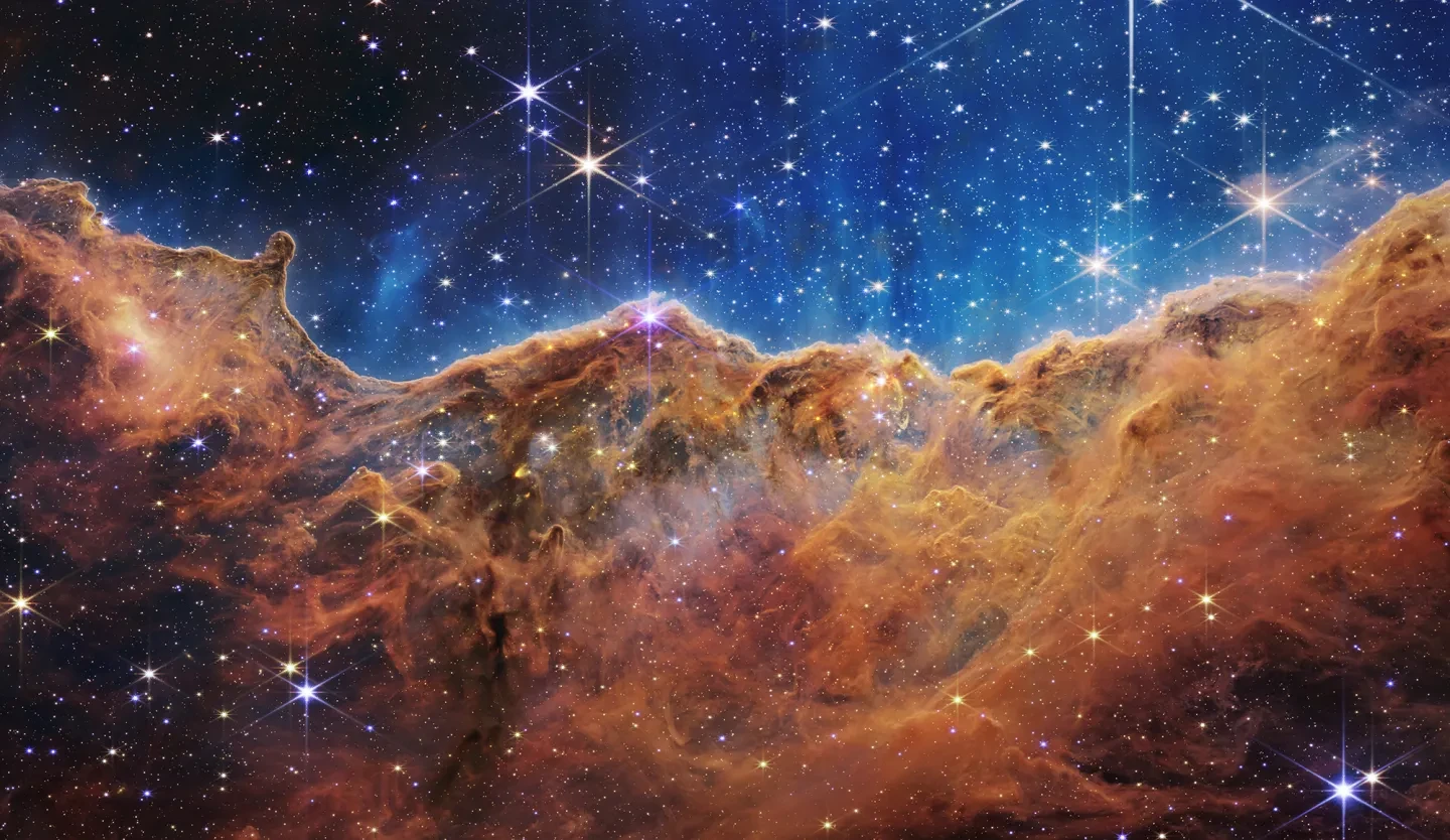 The image is divided horizontally by an undulating line between a cloudscape forming a nebula along the bottom portion and a comparatively clear upper portion. Speckled across both portions is a starfield, showing innumerable stars of many sizes. The smallest of these are small, distant, and faint points of light.