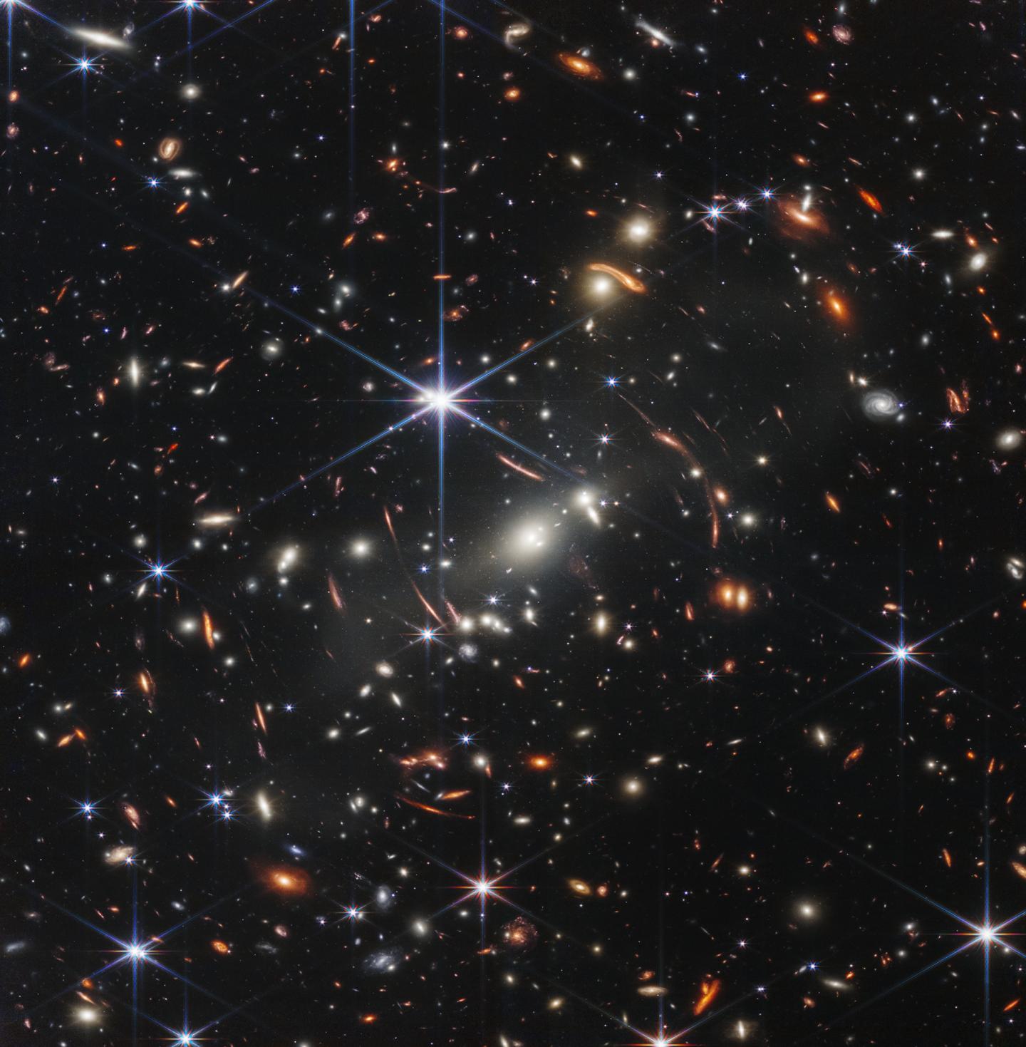 Thousands of small galaxies appear across this view. Their colors vary. Some are shades of orange, while others are white. Most appear as fuzzy ovals, but a few have distinct spiral arms. In front of the galaxies are several foreground stars. Most appear blue, and the bright stars have diffraction spikes, forming an eight-pointed star shape. There are also many thin, long, orange arcs that curve around the center of the image