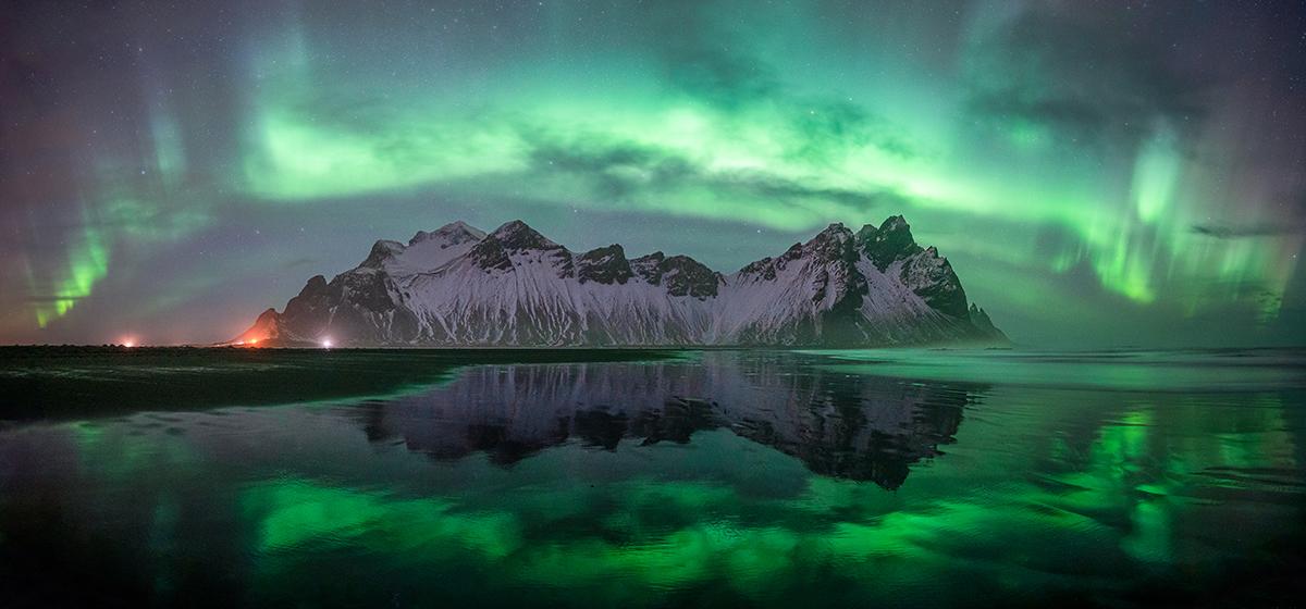 Green aurorae arcing over snowy mountains beside a still lake