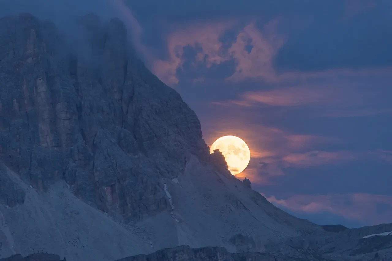 The Moon appears just above a snowy mountain, in a dark purple and pink sky