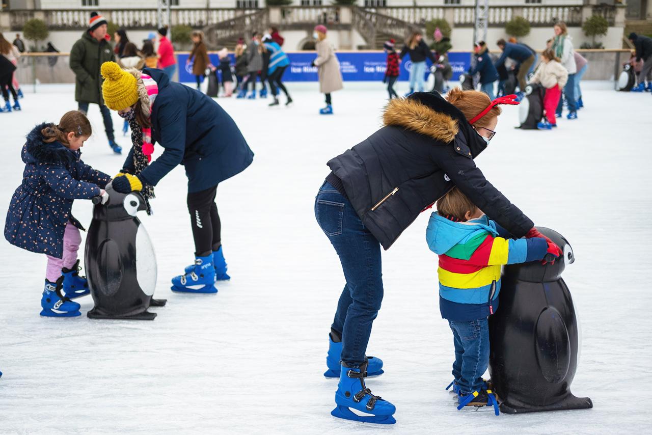 Two mothers and their children play on the Queen's House Ice Rink. They are using skate aids shaped like penguins to help balance