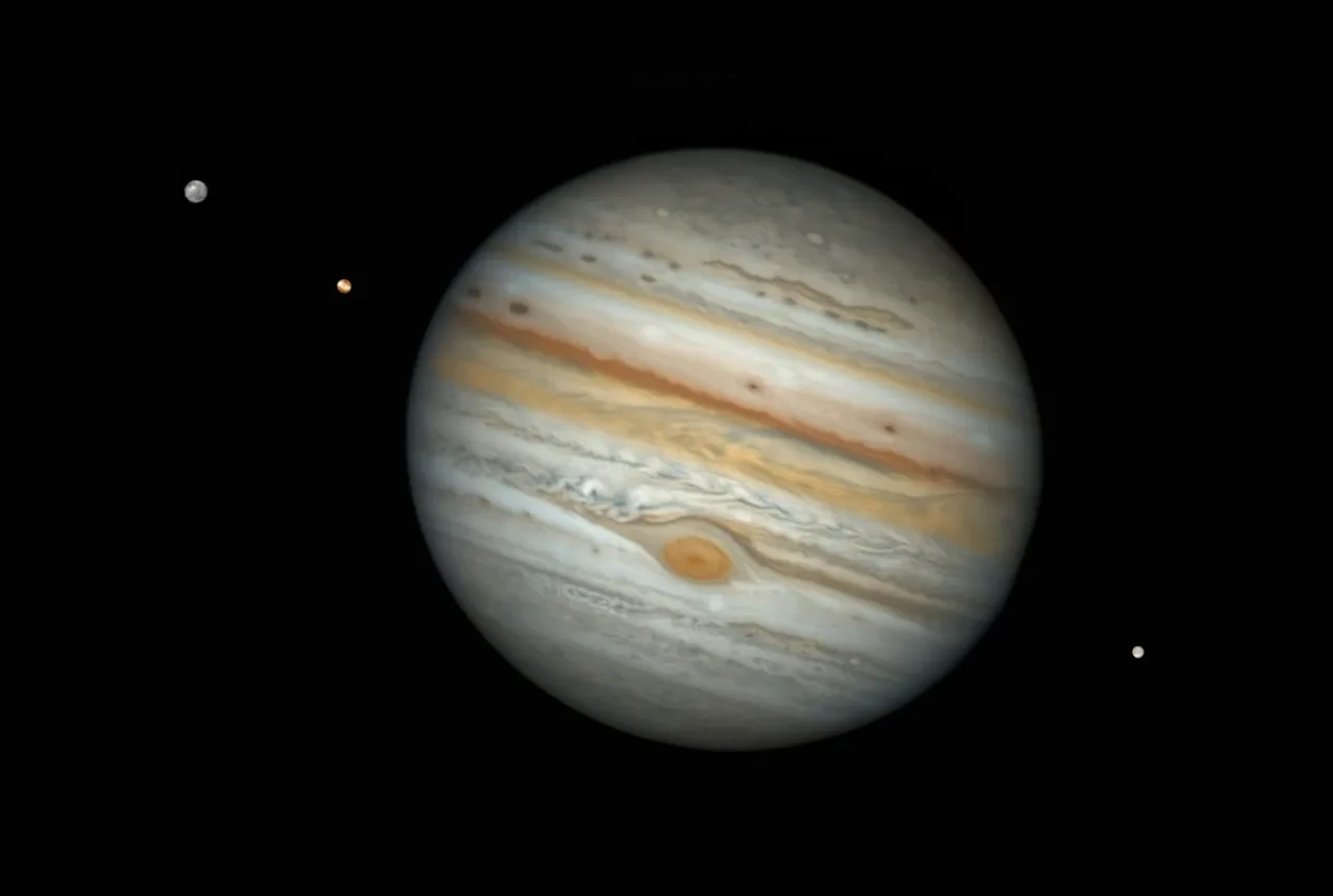 Image of Jupiter at a slight tilt, with three moons around it, two on a diagonal on the top left hand side and one on the bottom right hand side