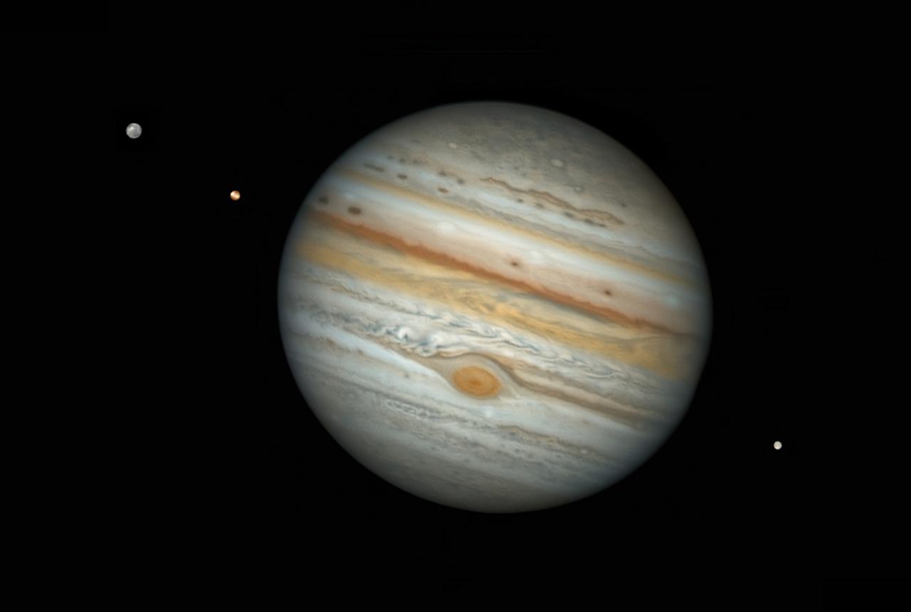 Image of Jupiter at a slight tilt, with three moons around it, two on a diagonal on the top left hand side and one on the bottom right hand side