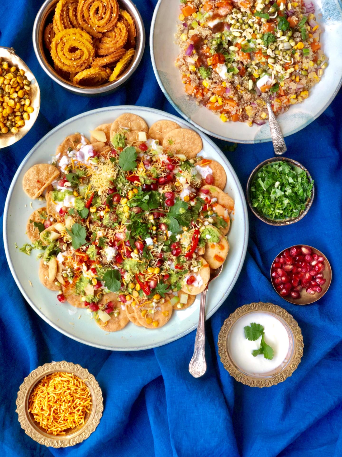 A display of different Indian food dishes laid out on a dark blue tablecloth. Large plates full of bhel and salad are accompanied with smaller dishes with chutneys and yoghurt