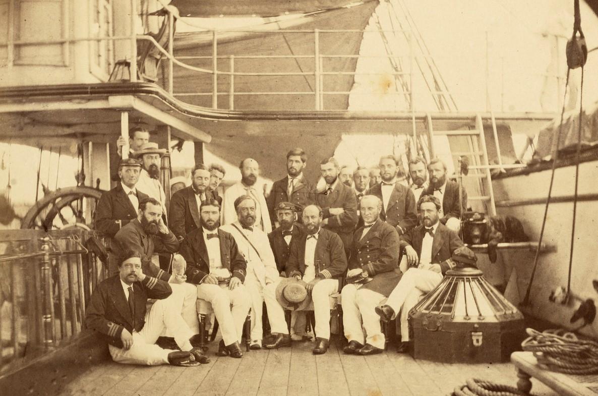 A sepia tone photograph taken in 1873 showing officers and scientists on board HMS Challenger. They are lined up in a group photo on the main deck of the ship, with the front row sat on chairs or the deck and the rear row standing behind
