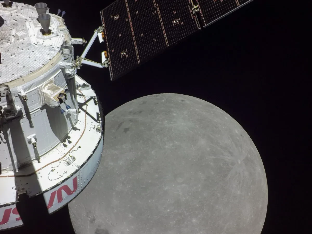 Image of white space capsule with solar panel extended in the left hand side of screen, with grey surface of the Moon in the bottom center