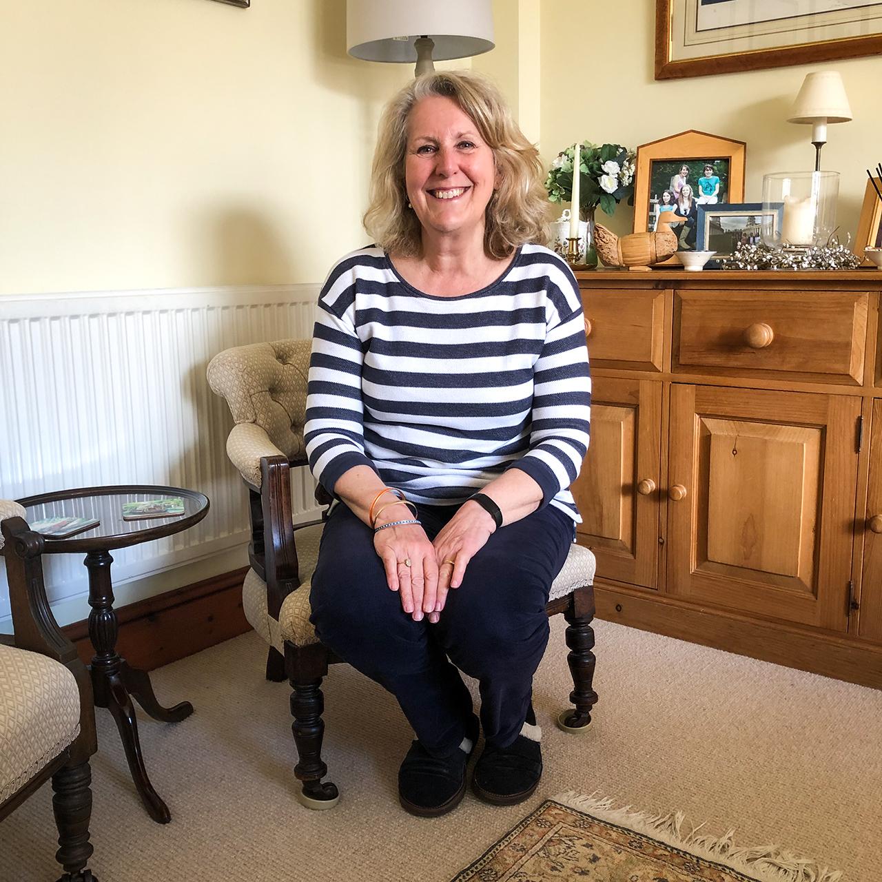 A woman in a striped top and dark trousers sits on a chair, smiling directly into the camera. She is at home, with family photos and candles on display on the dresser behind