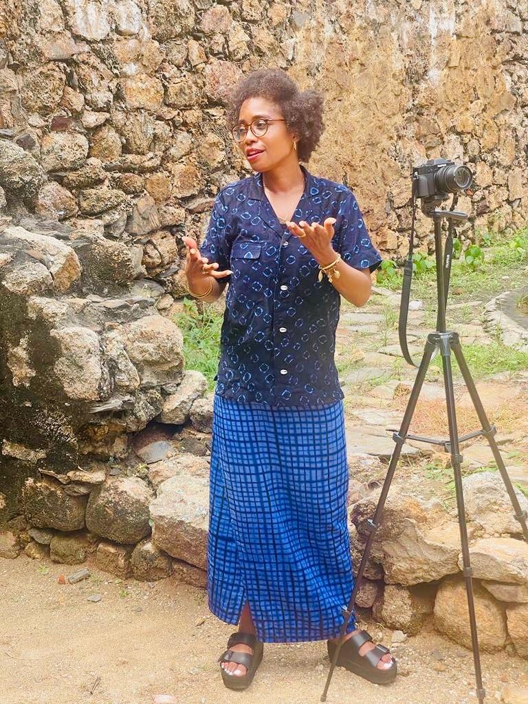 Nydia Swaby speaking to camera in front of a stone wall, a section of Fort Amsterdam, a former slave fort on the West Coast of Ghana 