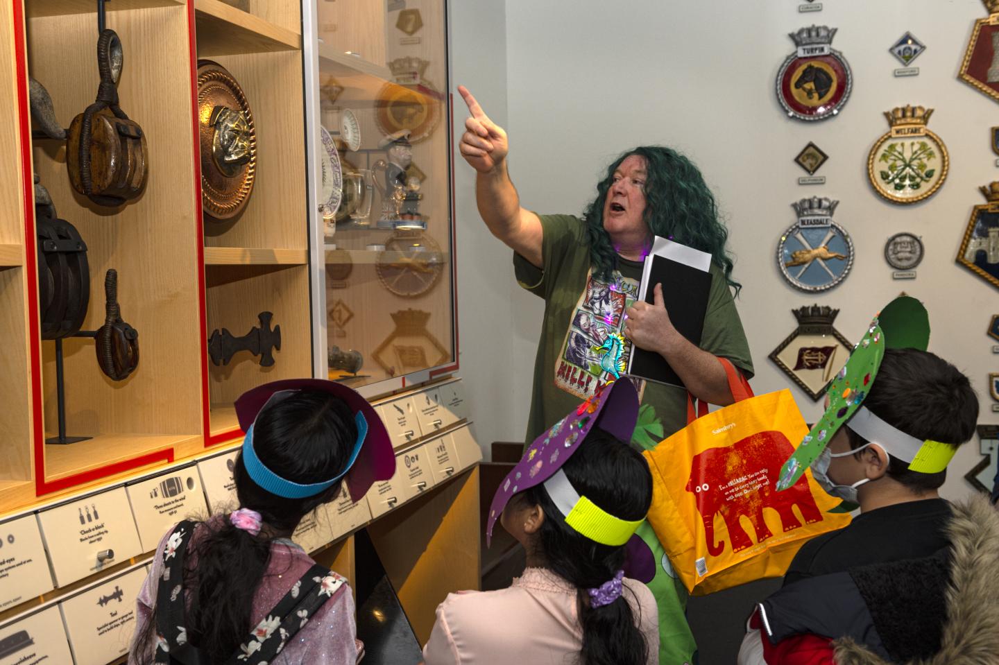 Children wearing handmade headdresses and listening to a talk by a visitor services assistant, who is pointing at some objects on display in a case