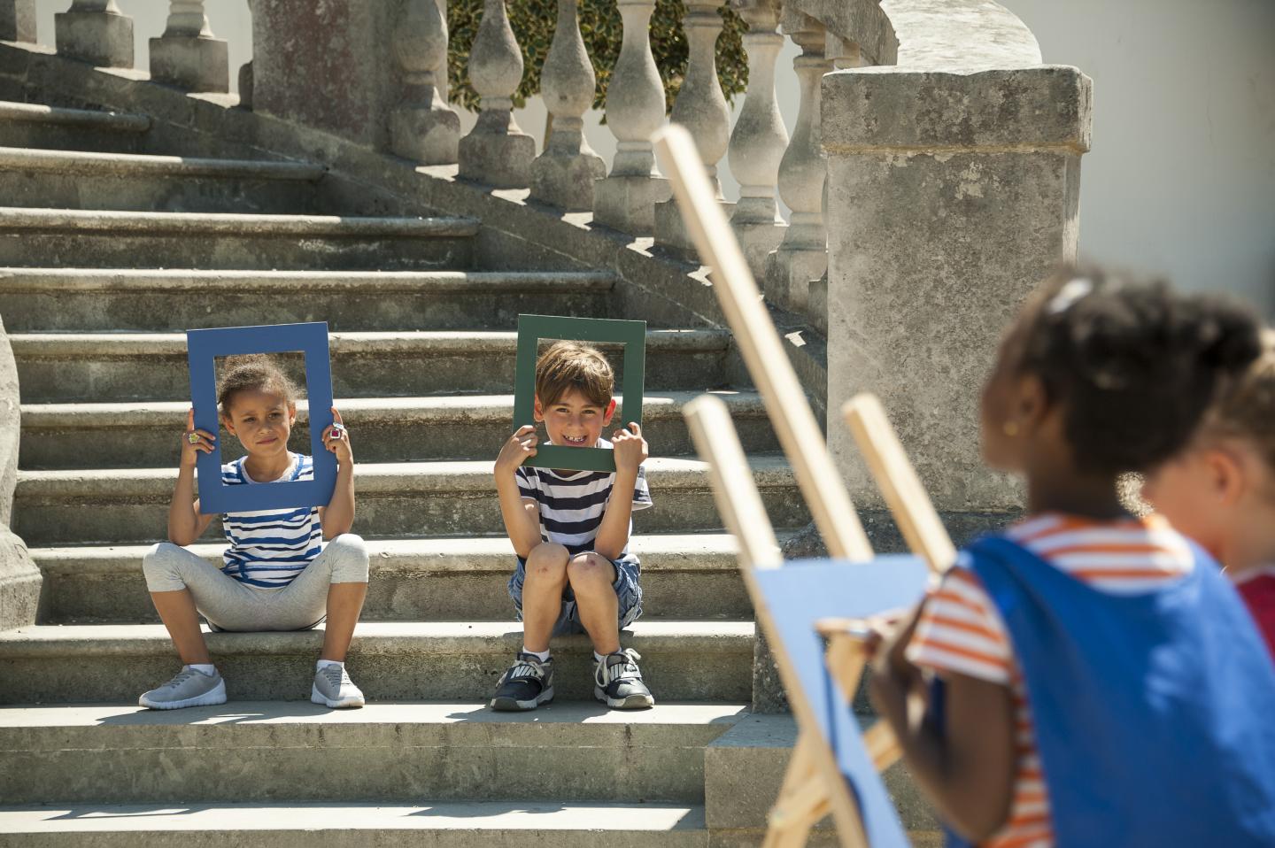 The image shows a child pretending to paint a portrait of two more children.