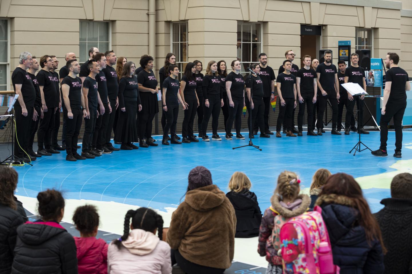 Image of choir of around 20 people wearing all black singing, with a choir master stood in front of them