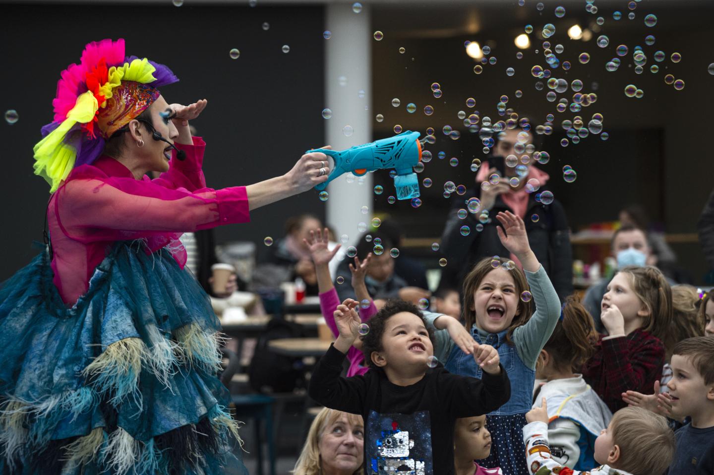 Performer dressed in garish pinks and yellows is using a bubble making device and children are happily reaching up to the bubbles and laughing
