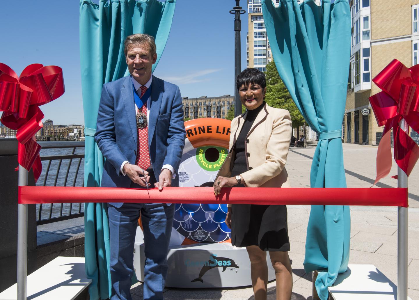 A man and a woman cut a ribbon tape to mark the installation of a new orange recycling bin on a seaside promenade