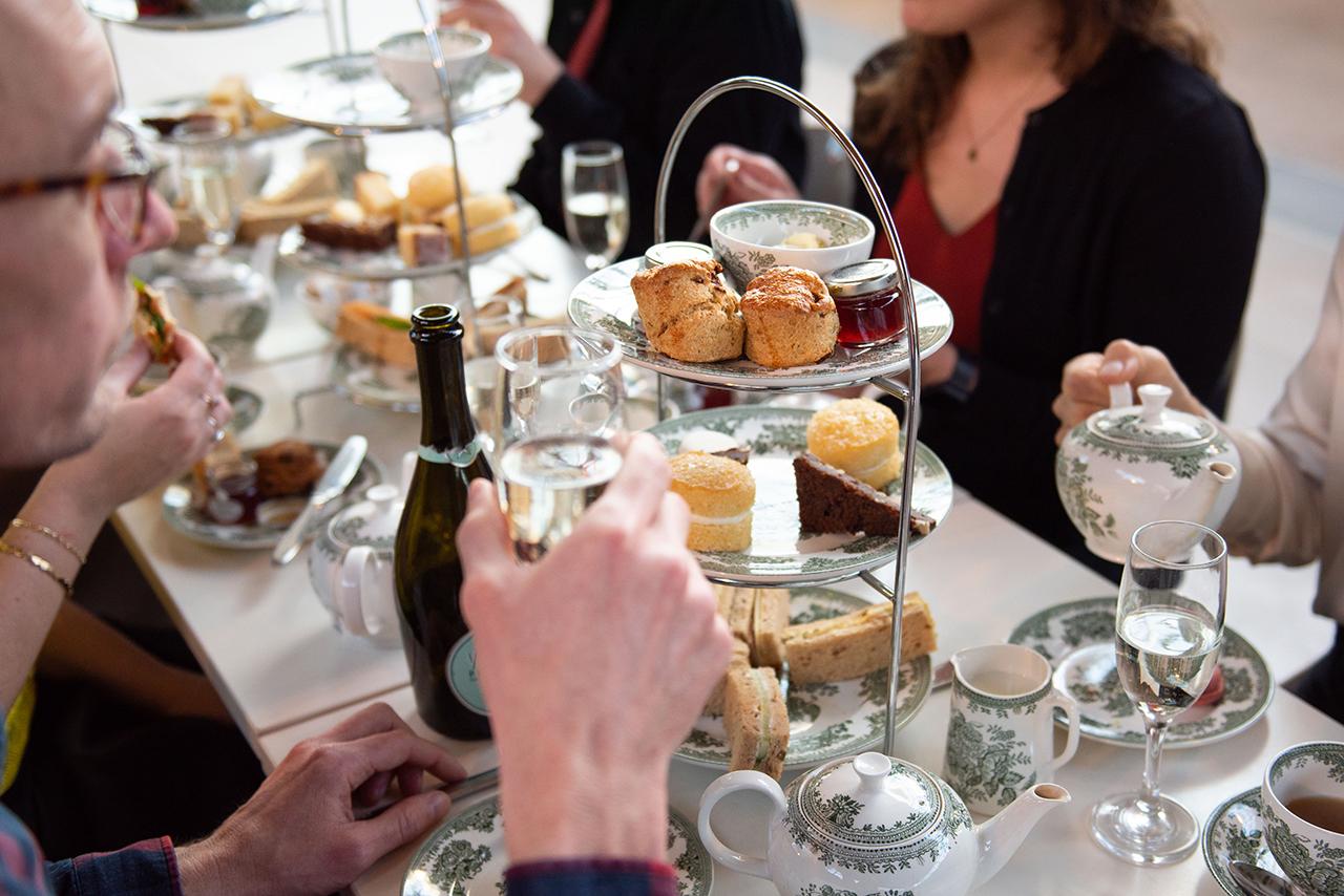 A table laid out for afternoon tea, with people gathered round a platter of sandwiches, scones and cakes, plus bottles of prosecco and pots of tea