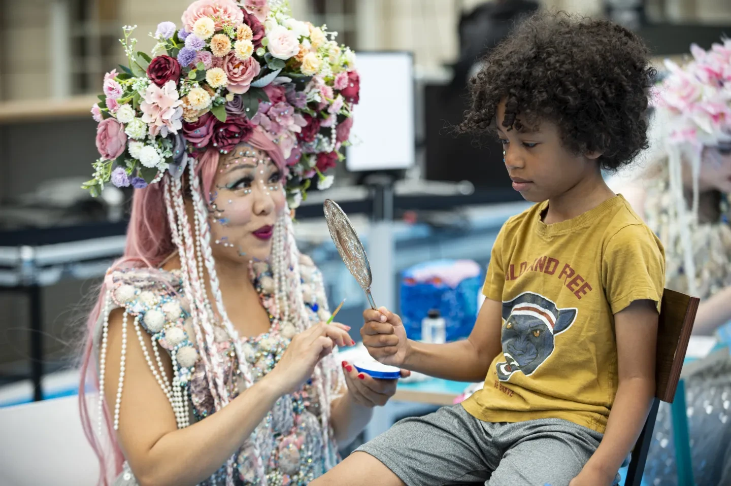 The image shows a child looking in a mirror next to a person dressed as a mermaid who has painted a design on the child's face.