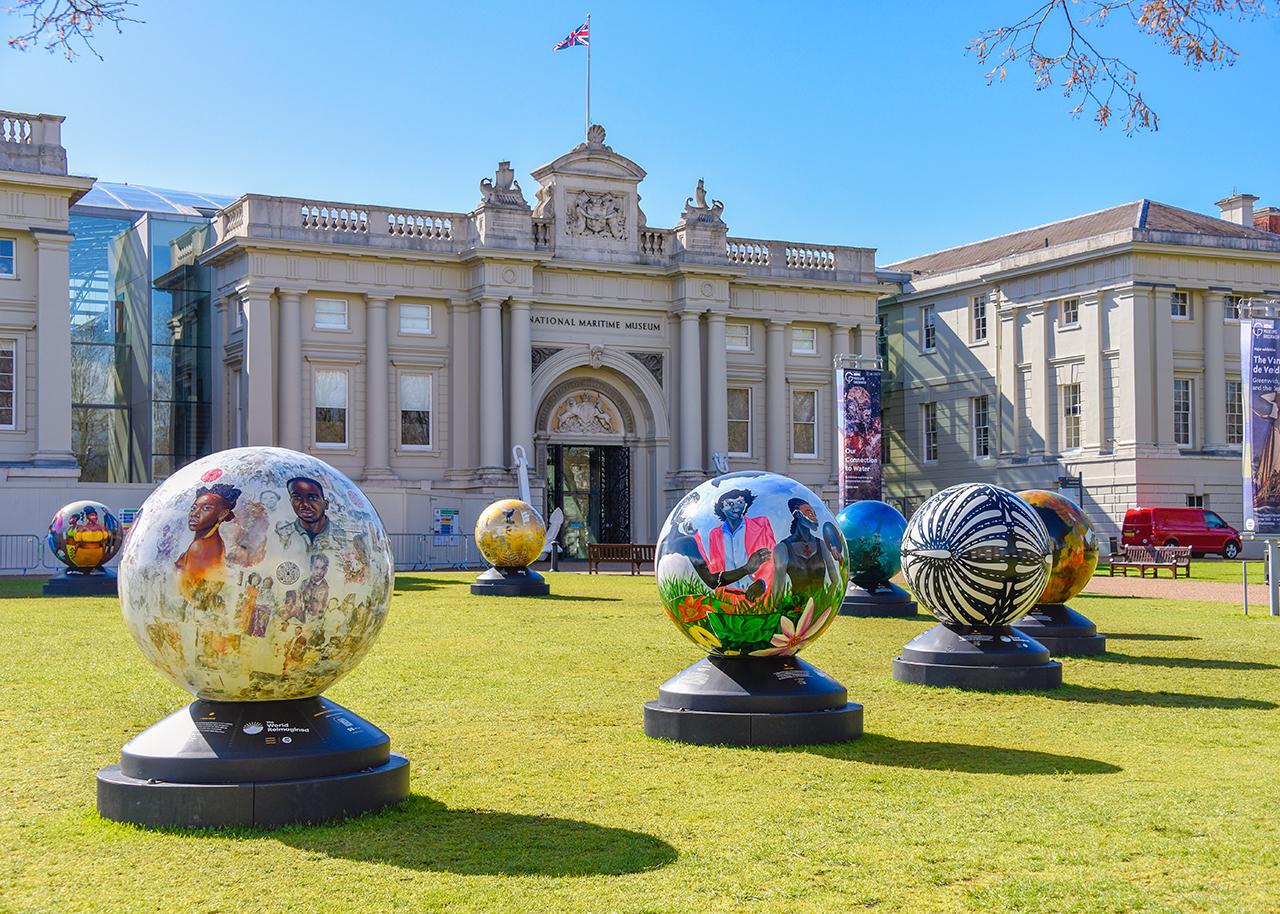 Photograph showing an art installation outside the National Maritime Museum. A series of artfully decorated globes are spread out on the lawns in front of the main building. Portraits of young Black people are visible on some, while another features a black and white striped pattern