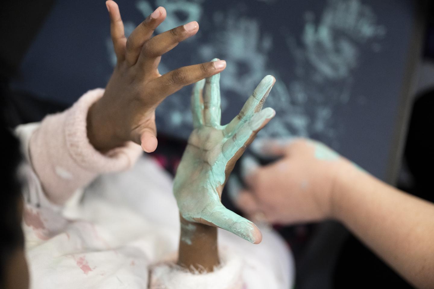 A child's hands photographed during an art workshop. The palm of their right hand is covered in light green paint, and they are feeling the wet paint with their left hand. In the background, a piece of paper with green handprints is just visible