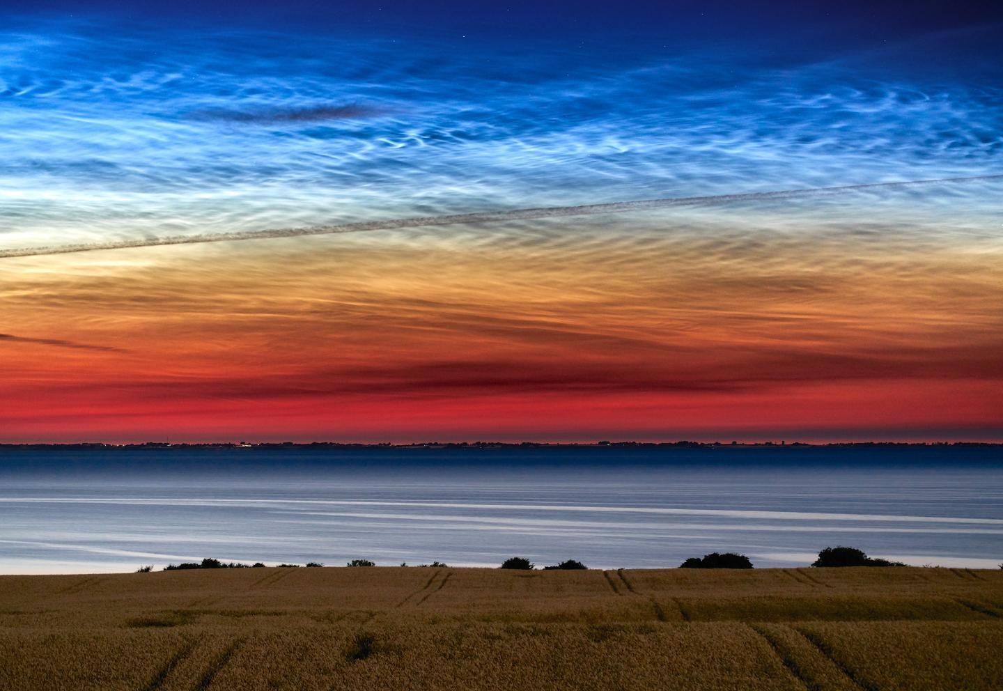 Image showing noctilucent clouds in sky above a golden field of wheat and the sea, the sky is red on the bottom, moving up to orange and through to the noctilucent clouds layer which is blue