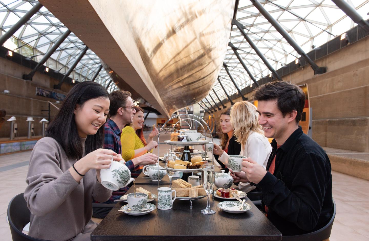 A group of diners enjoy afternoon tea beneath the historic ship Cutty Sark. The ship's hull can be seen 'floating' above them, supported by steel girders and a glass structure. On the table are cakes, sandwiches and fine china