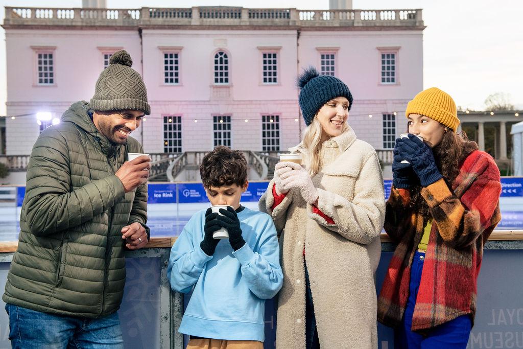 Family dressed in winter coats, hats and gloves having hot drinks from paper cups in front of the Queen's House Ice Rink, they are smiling at each other