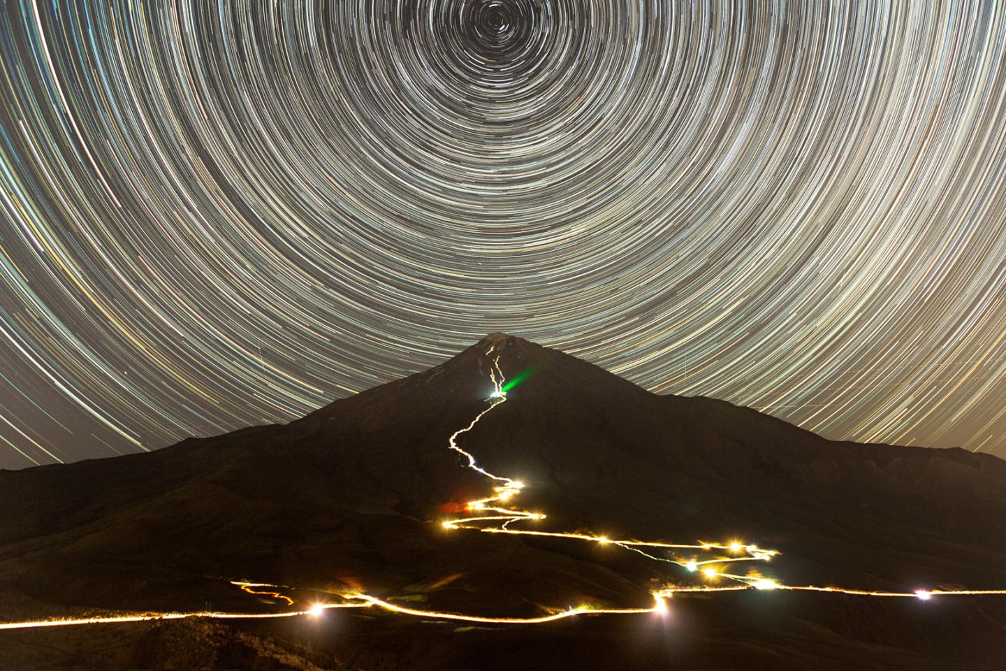 Image of a large mountain at night, with a road snaking up it illuminated in gold. In the sky is a semicircular star trail half cut off by the edges of the image in whites and yellows