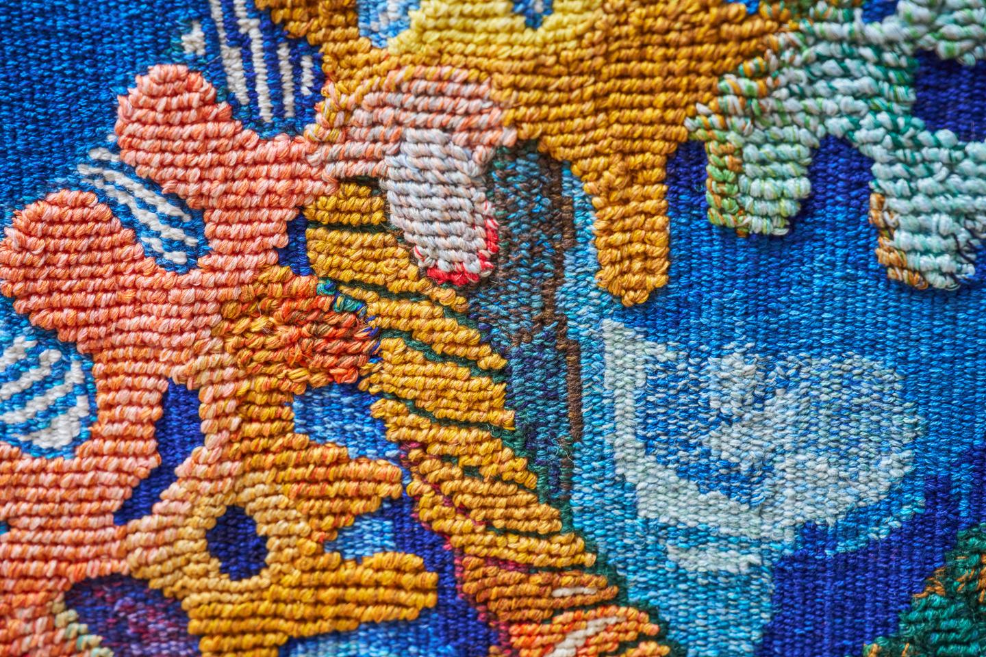 Close up image of the weaving of a tapestry, showcasing orange corals and a twisted yellow rope on a blue background