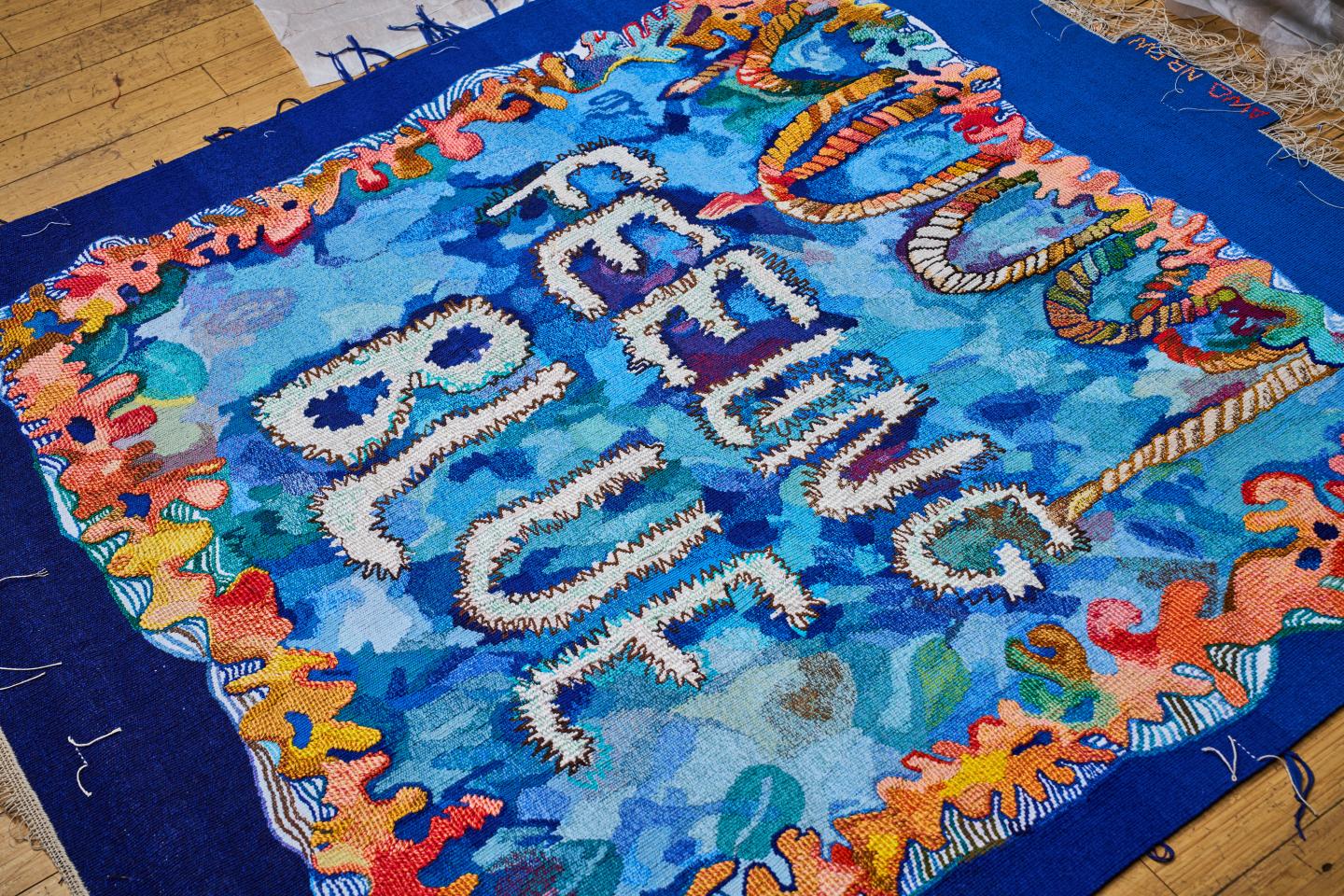 A blue tapestry is laid out on the floor, framed with colourful borders of coral and with the words 'Feeling Blue' woven in the centre