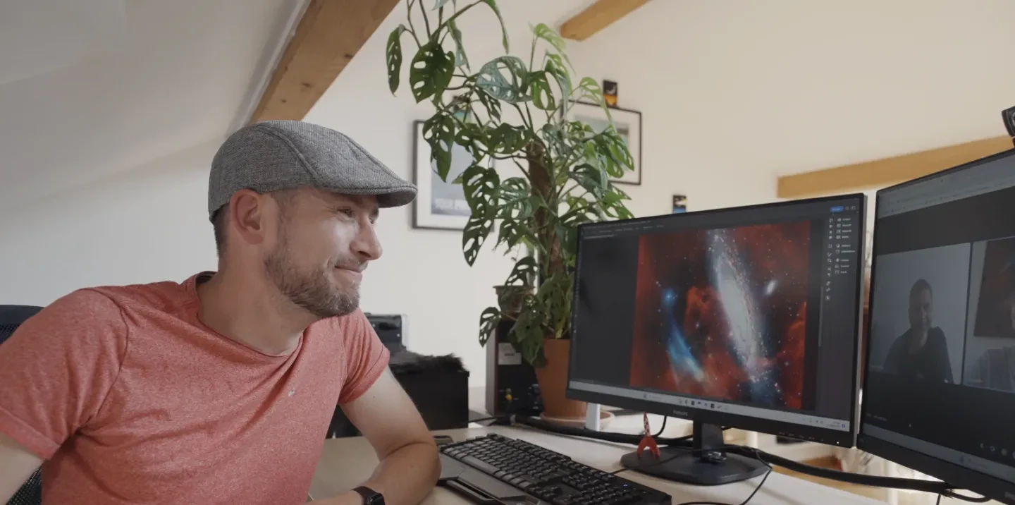 Image of man sitting at a computer with image of a galaxy with plasma arc on one screen and two men on Zoom on other screen