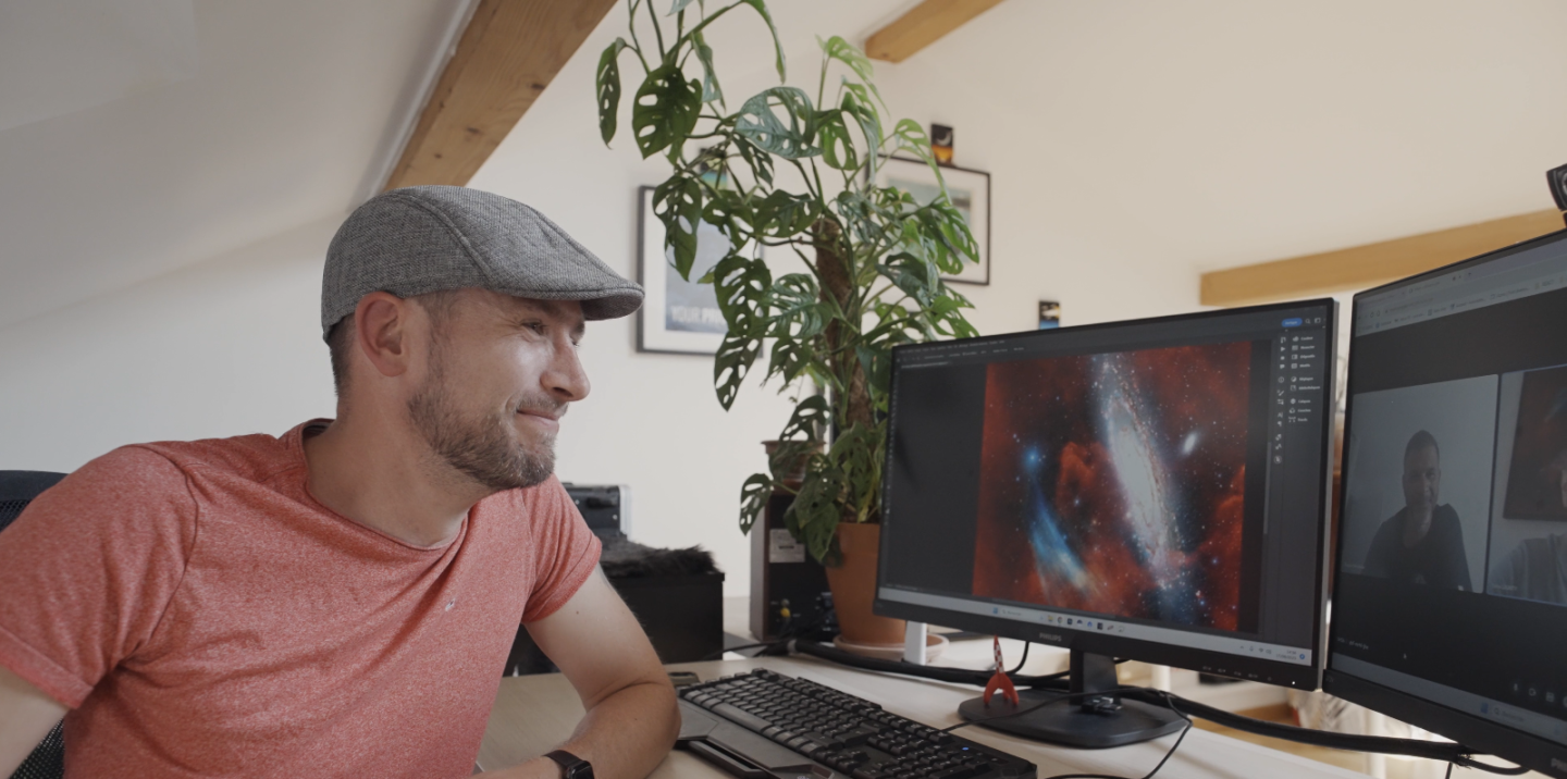 Image of man sitting at a computer with image of a galaxy with plasma arc on one screen and two men on Zoom on other screen