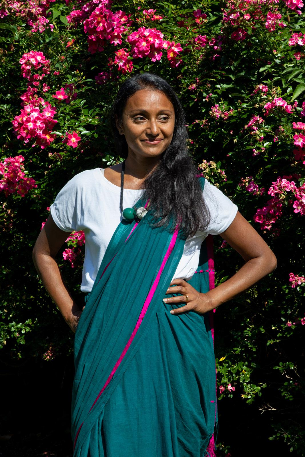 Photograph of a woman standing in front of a pink rose bush. She is wearing a blue sari draped across her left shoulder, and is standing with her hands on her hips, smiling and looking off to the left.