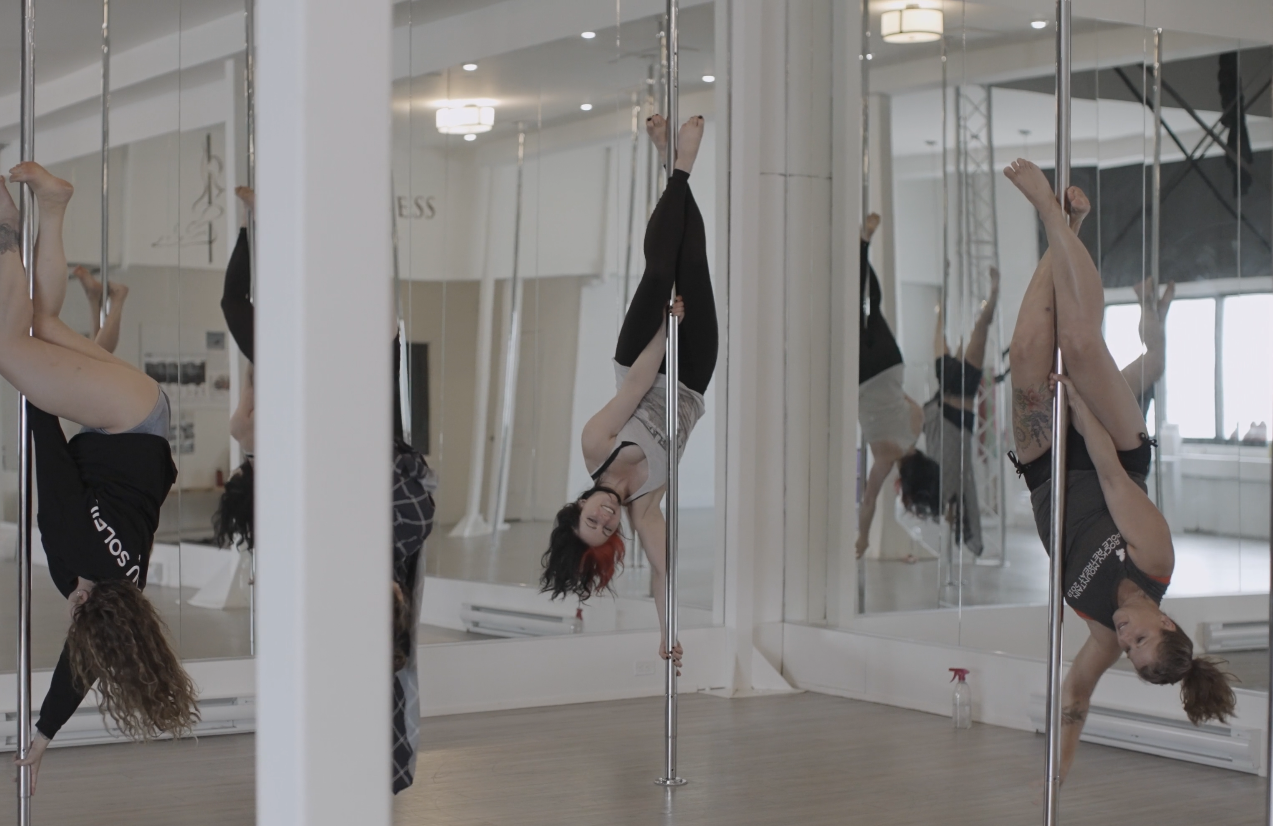Image of woman pole dancing, amongst two other women, upside down and spinning around in a pole dancing studio