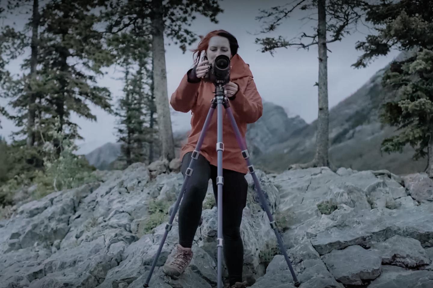 Image of woman wearing a red coat taking a photo using a camera on a tripod, with rocks, trees and mountains in the background