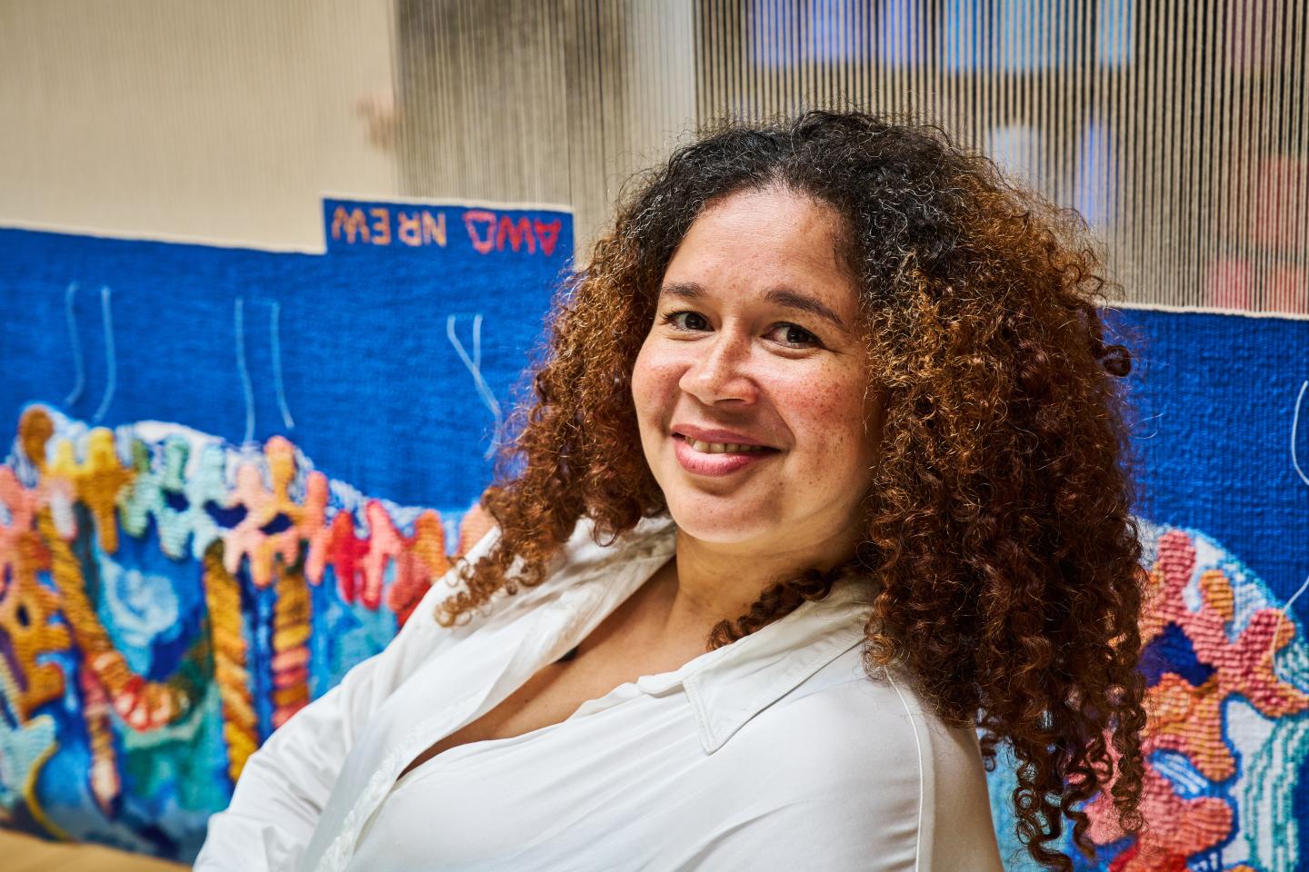 Alberta Whittle smiles at the camera with a blue coloured tapestry called Feeling Blue on a loom in the background