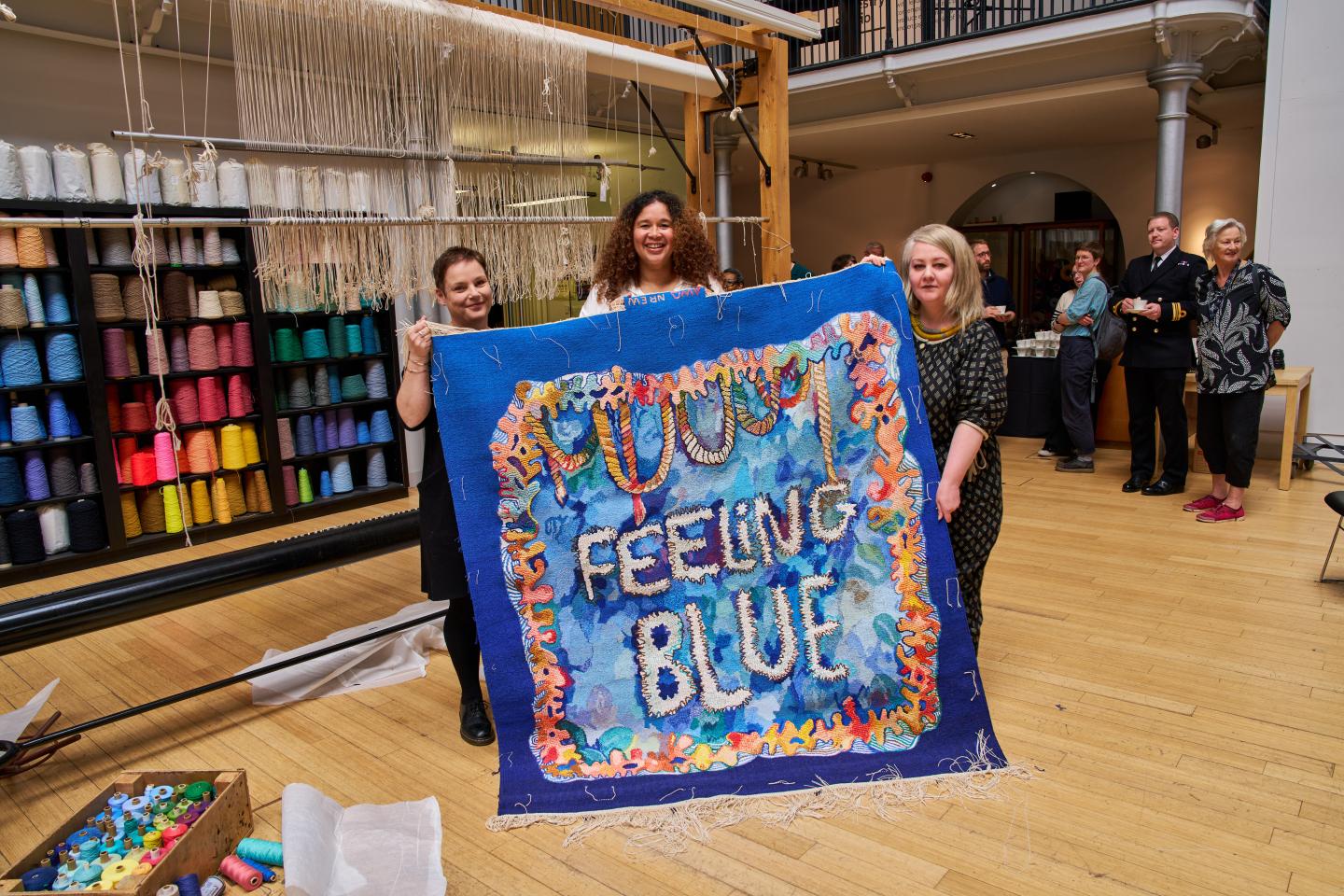 A group of weavers stand in front of a loom holding a blue tapestry with a colourful coral border and with the words 'Feeling Blue' woven in the centre