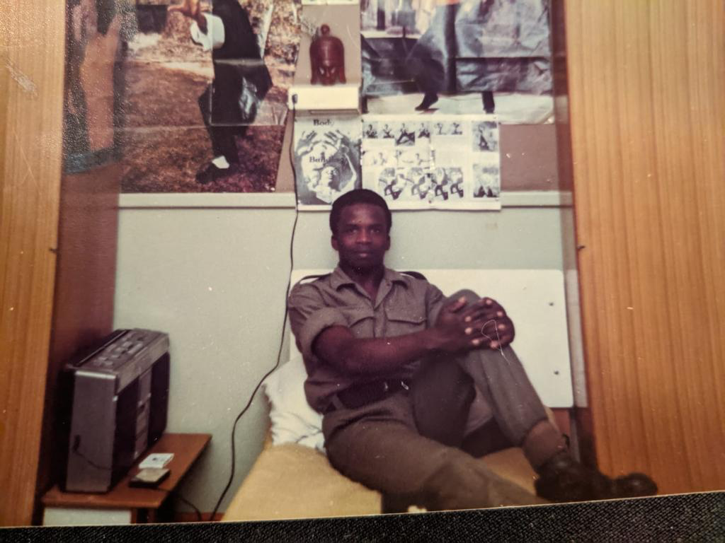 A black, male soldier sitting with radio beside him and photos and posters on the wall behind.
