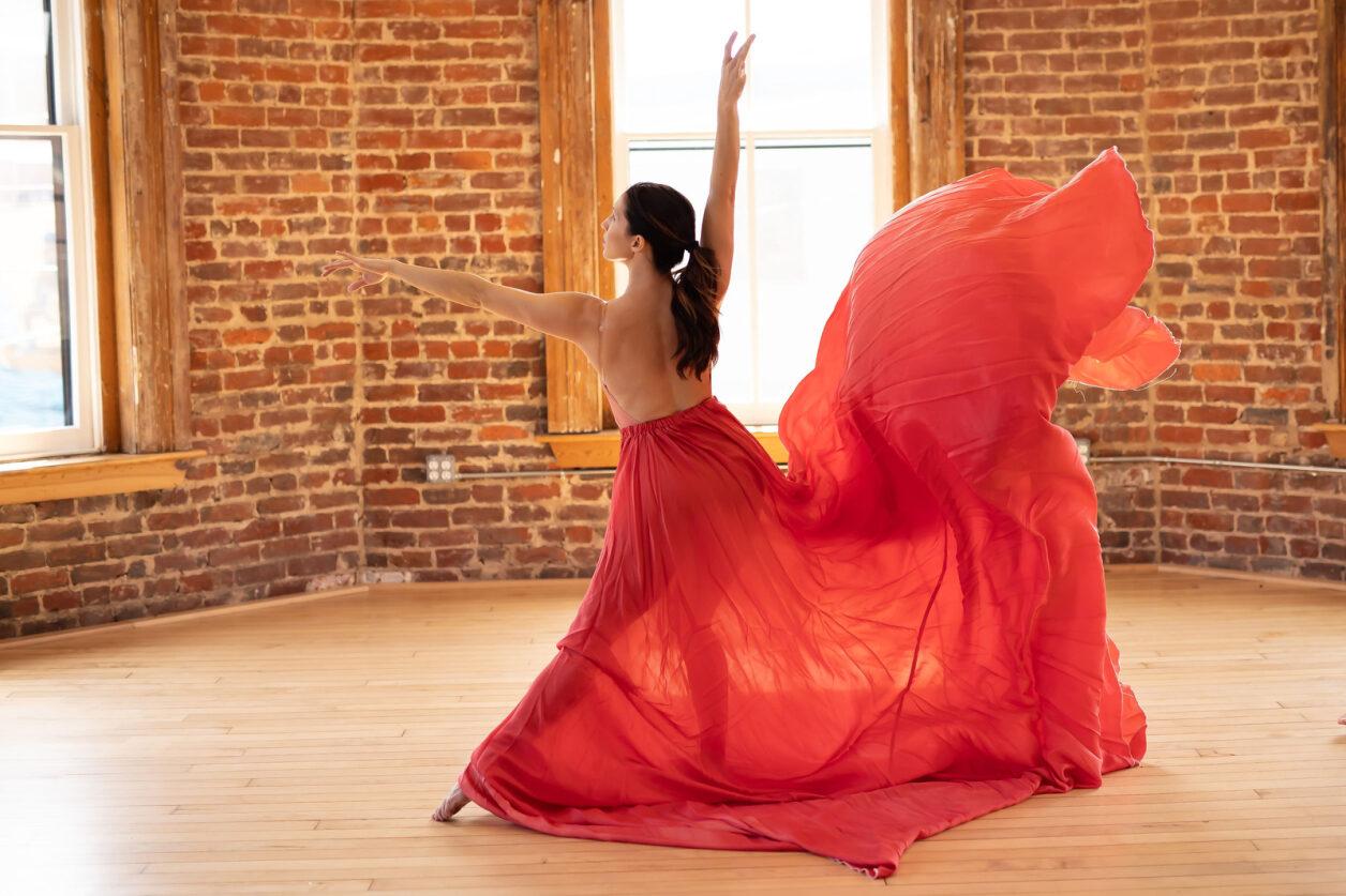 a woman in a dance pose with a floating red dress, the background is a bare wall.