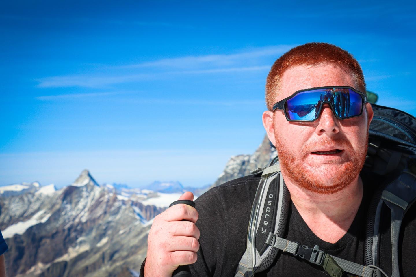 colour photo of a man with red hair and beard, with sky and mountains behind, as he is climbing.