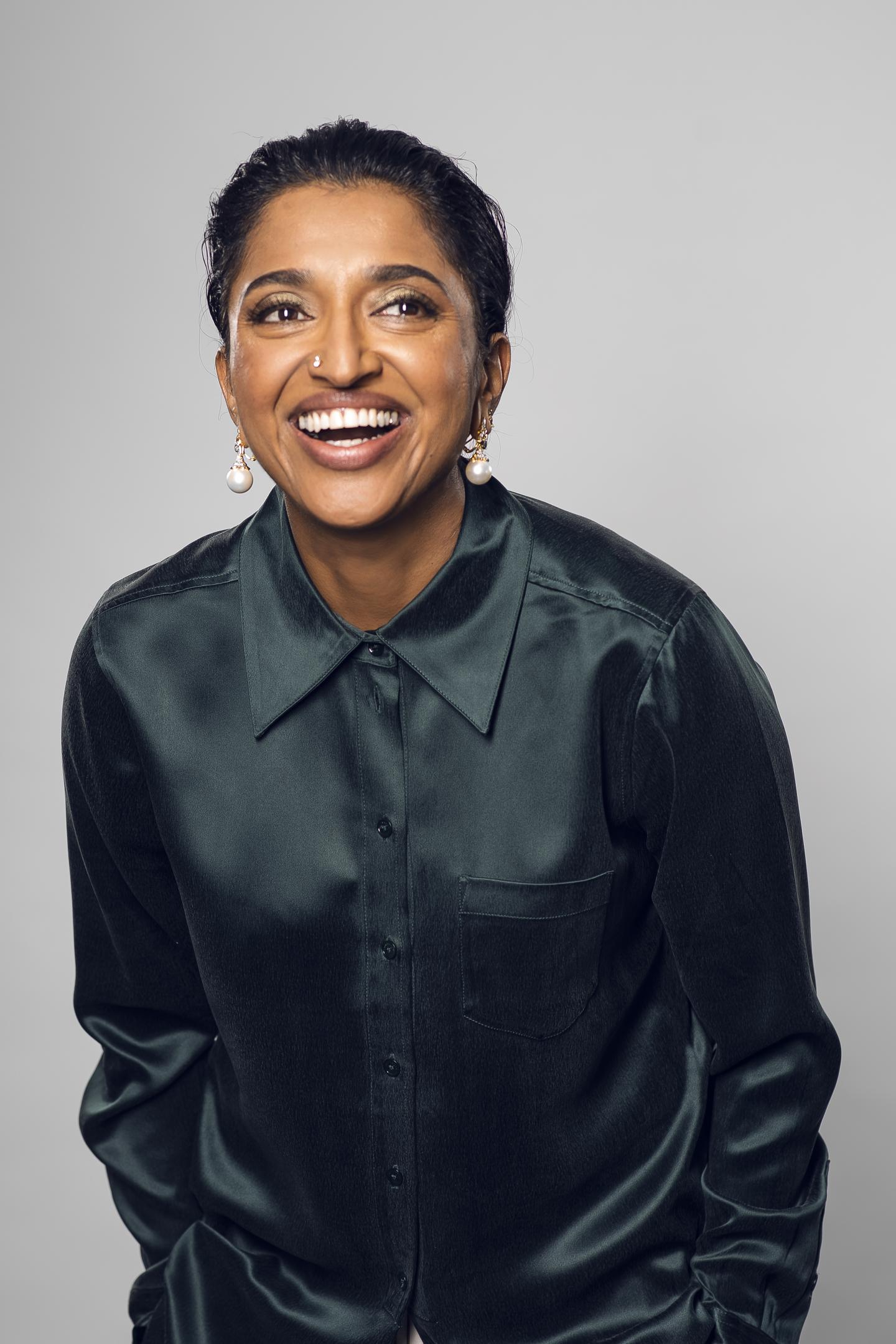 Image of comedian Sindhu Vee, wearing a black silk shirt and smiling