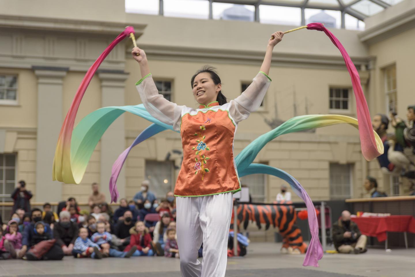 The image shows a woman performing a dance with ribbons in front of a crowd.