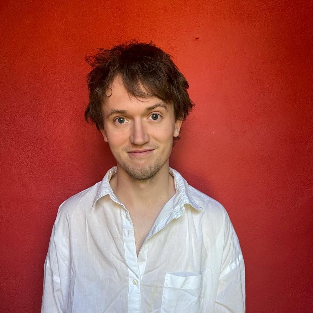 Image of comedian Daniel McKeon, who has short brown hair and is wearing a white shirt in front of a red background