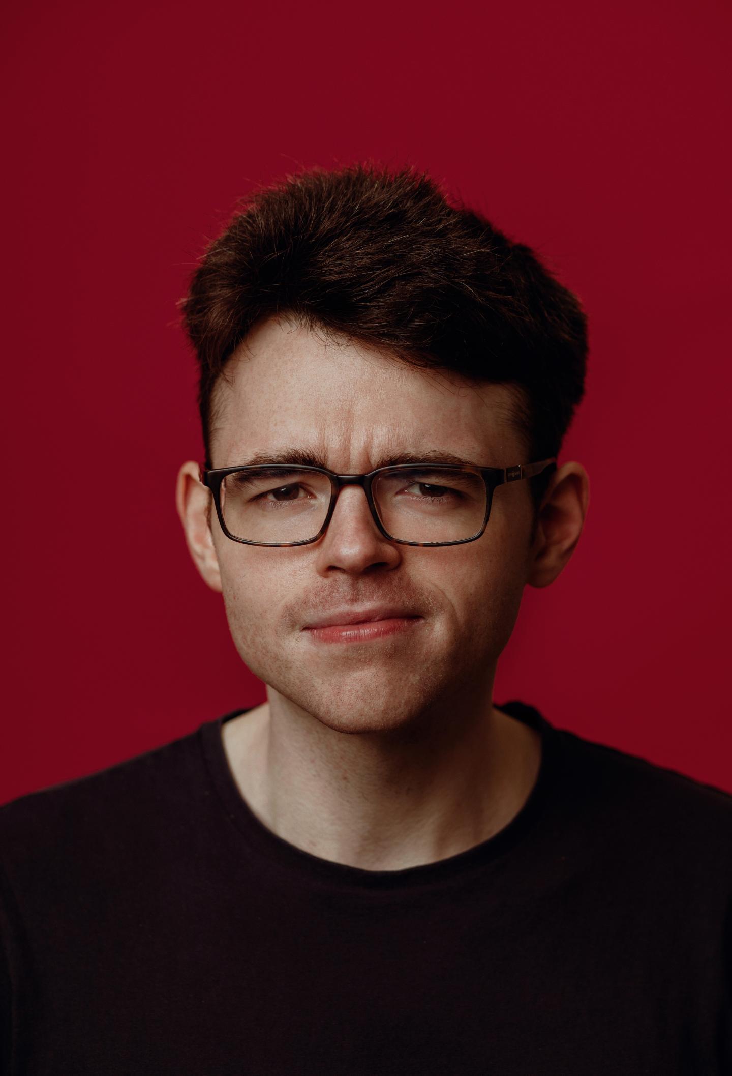 Image of comedian Roger O'Sullivan, against a red background wearing glasses with a side part in his hair