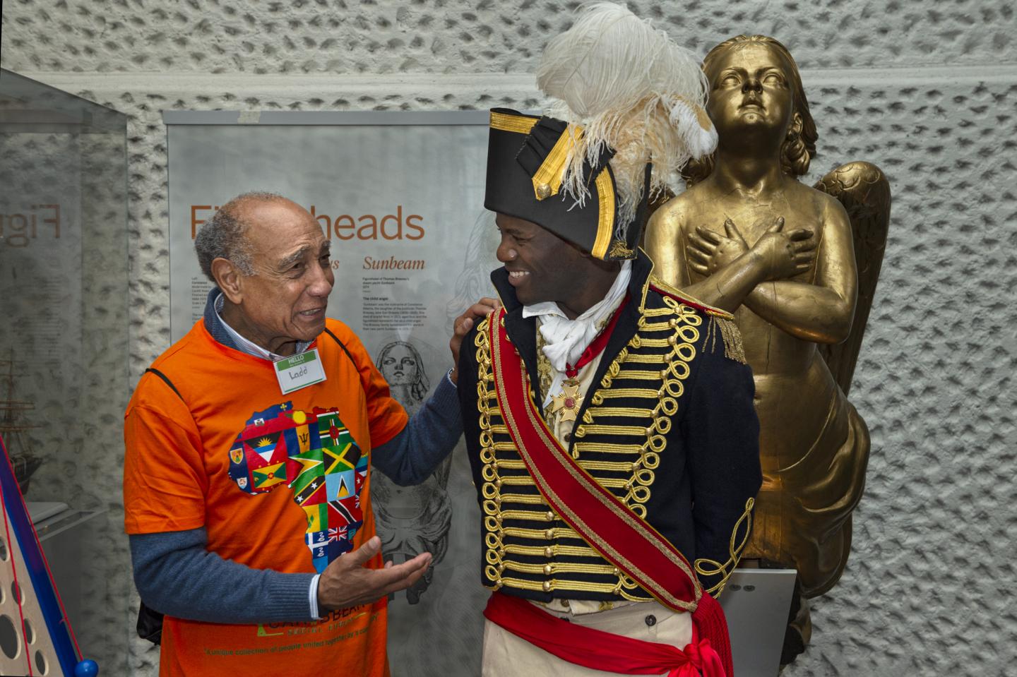 A man dressed in a historical uniform laughs with a man dressed normally in front of a ship figurehead